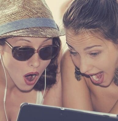 two women wearing hats and sunglasses look at a laptop screen